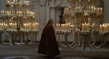 Movie still from “Fellini's Casanova” (1976), directed by Federico Fellini – A woman in a long brown coat standing in front of a group of lit candles; Wide shot, High angle