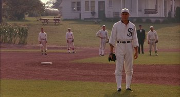 Movie still from “Field of Dreams” (1989), directed by Phil Alden Robinson – A baseball player is standing on a field with other baseball players in the background; Wide shot, Over the shoulder angle