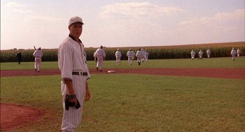 Movie still from “Field of Dreams” (1989), directed by Phil Alden Robinson – A baseball player standing on a baseball field with other baseball players in the background; Wide shot, Over the shoulder angle