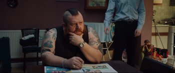 Movie still from “Fighting with My Family” (2019), directed by Stephen Merchant – A man sitting at a table with his hand under his chin; Medium shot, Over the shoulder angle