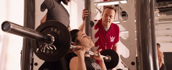 Movie still from “Fighting with My Family” (2019), directed by Stephen Merchant – A woman lifting a barbell in front of two men; Medium shot, Over the shoulder angle