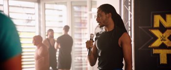 Movie still from “Fighting with My Family” (2019), directed by Stephen Merchant – A man holding a microphone while standing in a room; Medium shot, Over the shoulder angle
