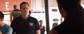 Movie still from “Fighting with My Family” (2019), directed by Stephen Merchant – A man standing in front of a group of people in a room; Medium shot, Over the shoulder angle