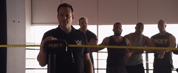 Movie still from “Fighting with My Family” (2019), directed by Stephen Merchant – A man standing next to two other men in a boxing ring; Medium shot, Over the shoulder angle