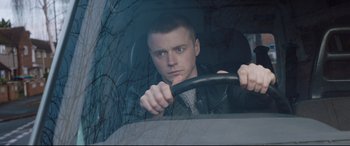 Movie still from “Fighting with My Family” (2019), directed by Stephen Merchant – A man driving a car in the rain with trees in the background; Close Up shot, Over the shoulder angle
