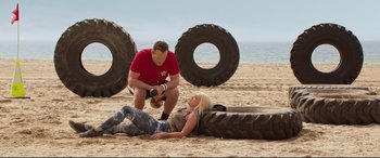 Movie still from “Fighting with My Family” (2019), directed by Stephen Merchant – A man kneeling down next to a woman laying on the beach; Wide shot, Over the shoulder angle