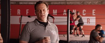 Movie still from “Fighting with My Family” (2019), directed by Stephen Merchant – A man holding a cup in his hand in front of an arena; Medium shot, Over the shoulder angle