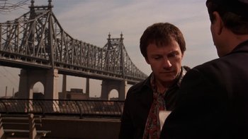 Movie still from “Fingers” (1978), directed by James Toback – A man standing next to a river with a bridge in the background; Medium shot, Over the shoulder angle