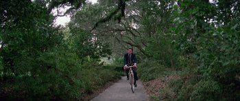 Movie still from “Firestarter” (1984), directed by Mark L. Lester – A man riding a bike down a path in the woods; Extreme Wide shot, High angle