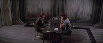Movie still from “Firestarter” (1984), directed by Mark L. Lester – Two people sitting at a table talking to each other; Wide shot, Over the shoulder angle
