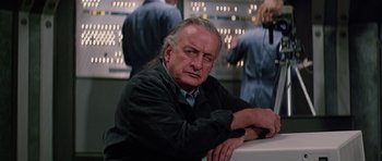 Movie still from “Firestarter” (1984), directed by Mark L. Lester – An older man sitting in front of a group of people; Close Up shot, Over the shoulder angle