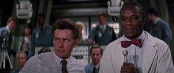 Movie still from “Firestarter” (1984), directed by Mark L. Lester – Two men are sitting next to each other in front of other men; Medium shot, Low angle