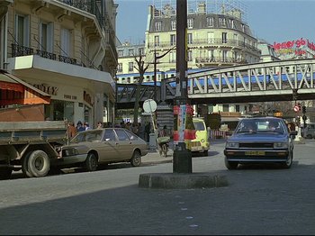 Movie still from “First Name: Carmen” (1983), directed by Jean-Luc Godard – A busy street with cars parked on the side of the road; Extreme Wide shot, High angle