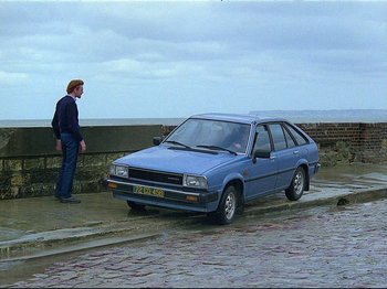 Movie still from “First Name: Carmen” (1983), directed by Jean-Luc Godard – A man standing next to a parked blue car; Wide shot, High angle
