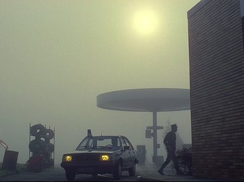 Movie still from “First Name: Carmen” (1983), directed by Jean-Luc Godard – A man walking down a street next to a gas station; Extreme Wide shot, Low angle