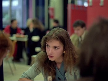 Movie still from “First Name: Carmen” (1983), directed by Jean-Luc Godard – A woman sitting at a table in front of a group of people; Medium shot, Over the shoulder angle
