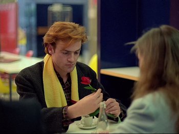 Movie still from “First Name: Carmen” (1983), directed by Jean-Luc Godard – A young man holding a red rose while sitting at a table with a woman; Medium shot, Over the shoulder angle
