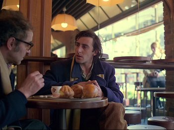 Movie still from “First Name: Carmen” (1983), directed by Jean-Luc Godard – Two men sitting at a table with a plate of food; Medium shot, Over the shoulder angle