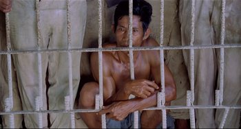 Movie still from “Fitzcarraldo” (1982), directed by Werner Herzog – A shirtless man sitting in a jail cell with his arms crossed; Medium shot, High angle