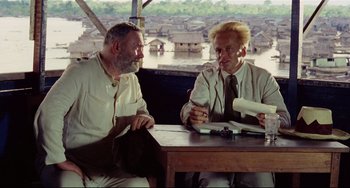 Movie still from “Fitzcarraldo” (1982), directed by Werner Herzog – Two men are sitting at a table and one of the men is holding papers; Medium shot, Over the shoulder angle