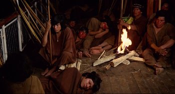 Movie still from “Fitzcarraldo” (1982), directed by Werner Herzog – A group of young people in native dress sitting around a fire; Wide shot, High angle
