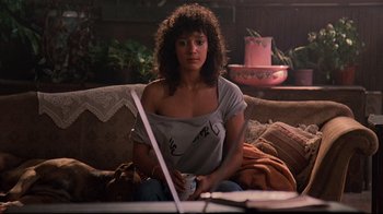 Movie still from “Flashdance” (1983), directed by Adrian Lyne – A woman sitting on a couch looking at a laptop computer; Medium shot, Over the shoulder angle