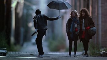 Movie still from “Flashdance” (1983), directed by Adrian Lyne – A man holding an umbrella while standing next to two women; Wide shot, Low angle