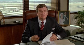 Movie still from “Fletch” (1985), directed by Michael Ritchie – A man in a suit and tie sitting at a desk; Medium shot, Over the shoulder angle