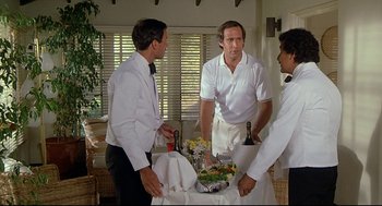 Movie still from “Fletch” (1985), directed by Michael Ritchie – A group of men standing around a table with food on it; Medium shot, High angle