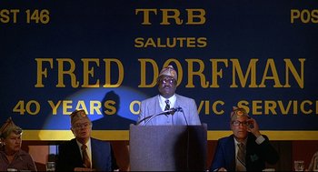 Movie still from “Fletch” (1985), directed by Michael Ritchie – A man is giving a speech at a podium; Medium shot, Low angle