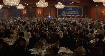 Movie still from “Fletch” (1985), directed by Michael Ritchie – A crowd of people sitting at tables in front of a microphone; Extreme Wide shot, High angle