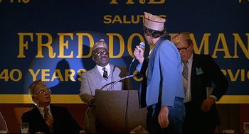 Movie still from “Fletch” (1985), directed by Michael Ritchie – A man is speaking at a podium while another man is wearing a hat; Medium shot, Low angle
