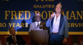 Movie still from “Fletch” (1985), directed by Michael Ritchie – A man is speaking at a podium while another man is holding a microphone; Medium shot, Low angle