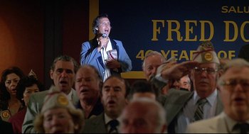 Movie still from “Fletch” (1985), directed by Michael Ritchie – A man in a blue jacket is speaking into a microphone in front of a group of people; Medium shot, Low angle