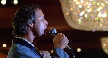 Movie still from “Fletch” (1985), directed by Michael Ritchie – A man is holding a microphone up to his mouth; Close Up shot, Low angle