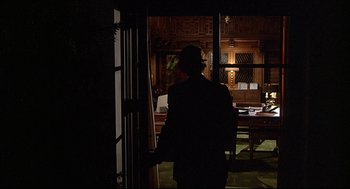 Movie still from “Fletch” (1985), directed by Michael Ritchie – A man standing in a room with a backpack; Wide shot, Over the shoulder angle