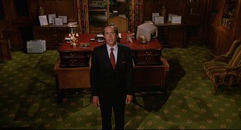 Movie still from “Fletch” (1985), directed by Michael Ritchie – A man in a suit standing in front of a desk in a room; Medium shot, Low angle