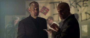 Movie still from “Flickering Lights” (2000), directed by Anders Thomas Jensen – A man in a suit and tie holding a cell phone; Medium shot, Low angle