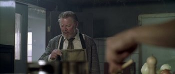 Movie still from “Flickering Lights” (2000), directed by Anders Thomas Jensen – An older man wearing an apron in a kitchen; Medium shot, Over the shoulder angle