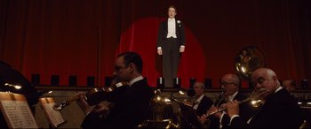 Movie still from “Florence Foster Jenkins” (2016), directed by Stephen Frears – A man in a tuxedo standing on a stage with other musicians; Wide shot, Low angle
