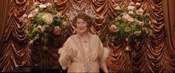 Movie still from “Florence Foster Jenkins” (2016), directed by Stephen Frears – An older woman wearing a tiara and holding a thumbs up; Medium shot, Over the shoulder angle