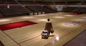Movie still from “Flubber” (1997), directed by Les Mayfield – A man in a brown jacket is pulling a luggage cart in an arena; Extreme Wide shot, High angle