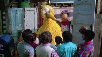 Movie still from “Follow That Bird” (1985), directed by Ken Kwapis – A group of people standing around a big yellow bird; Wide shot, Over the shoulder angle