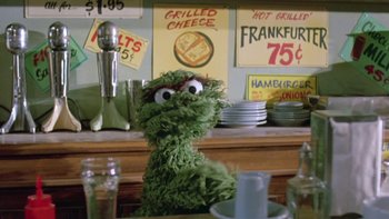 Movie still from “Follow That Bird” (1985), directed by Ken Kwapis – A green stuffed animal sitting on top of a table; Medium shot, Low angle