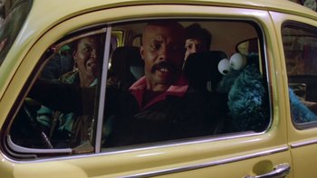 Movie still from “Follow That Bird” (1985), directed by Ken Kwapis – Three people in a yellow car with a sesame street character in the back seat; Medium shot, Over the shoulder angle