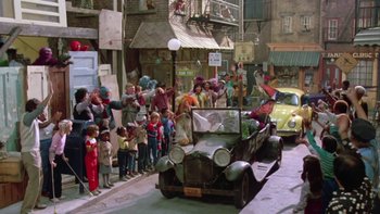 Movie still from “Follow That Bird” (1985), directed by Ken Kwapis – An old fashioned car driving down a street with a crowd of people watching it; Wide shot, High angle