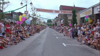 Movie still from “Follow That Bird” (1985), directed by Ken Kwapis – A crowd of people on a street under a banner; Extreme Wide shot, High angle
