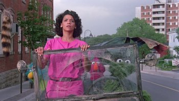 Movie still from “Follow That Bird” (1985), directed by Ken Kwapis – A woman standing next to a window with a sesame street character behind it; Medium shot, Low angle