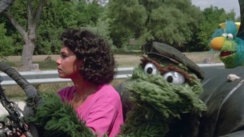 Movie still from “Follow That Bird” (1985), directed by Ken Kwapis – A woman sitting next to a person wearing a green monster costume; Medium shot, Low angle