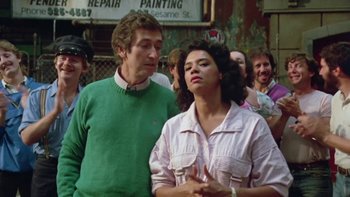 Movie still from “Follow That Bird” (1985), directed by Ken Kwapis – A man and a woman standing next to each other in front of a group of people; Close Up shot, Over the shoulder angle